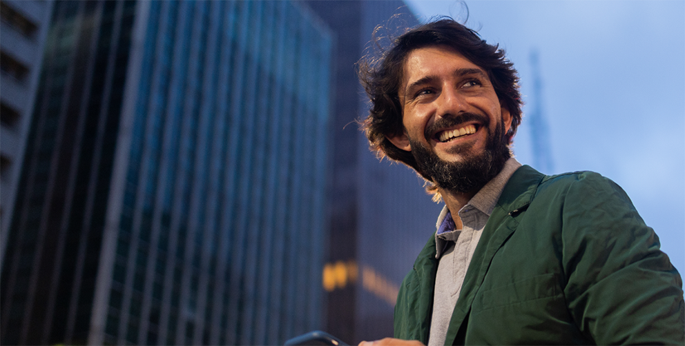 Smiling man in a green jacket holding a phone, standing in an urban cityscape with tall buildings in the background.